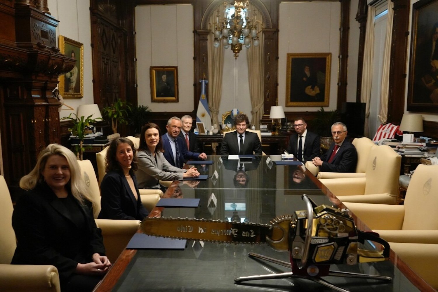 A group of people seated around a table, including RFK Jr. and Javier Milei, with a chainsaw on the table in front of them.